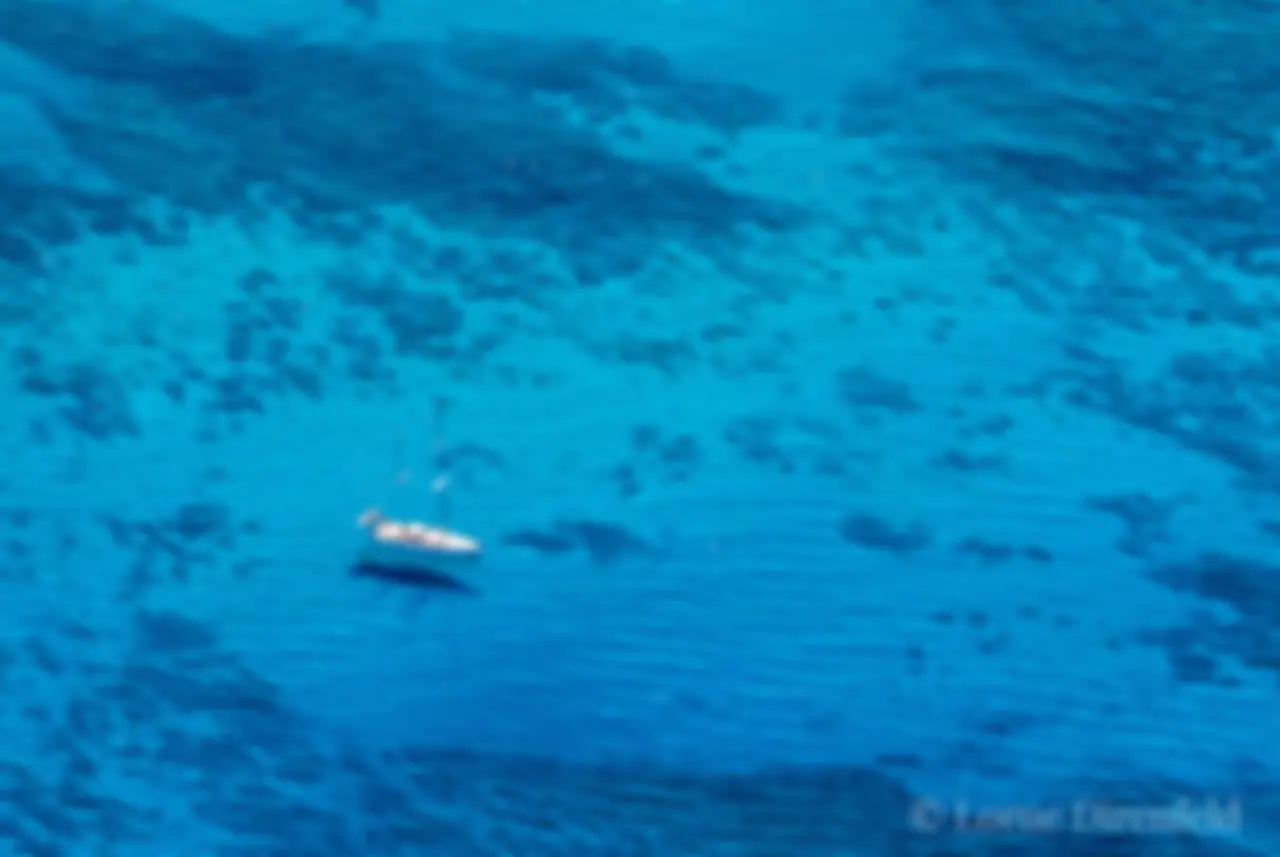 The surfers appear as small dots on the ocean as a sail boat hangs out in the morning in the shimmering blue waters off Waikiki . . .