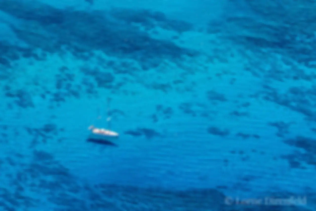 The surfers appear as small dots on the ocean as a sail boat hangs out in the morning in the shimmering blue waters off Waikiki . . .